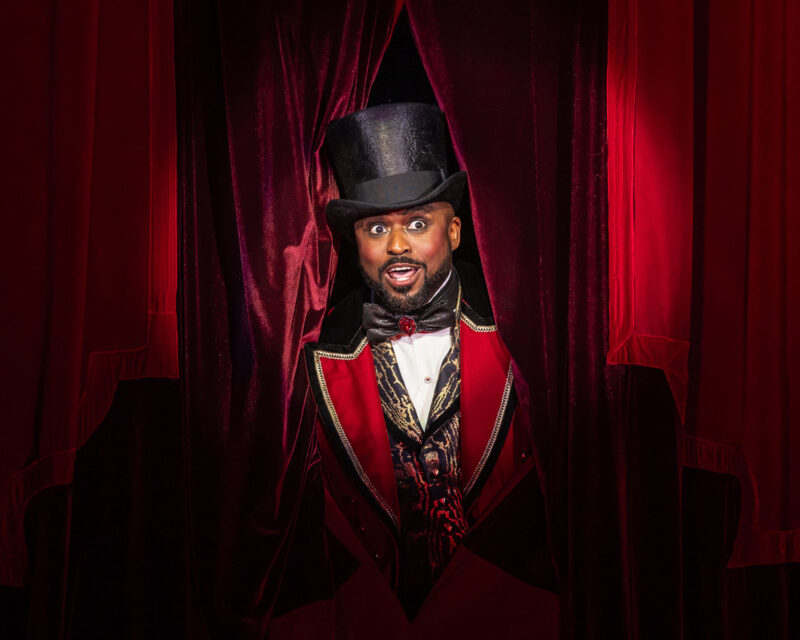Il maestro di cerimonie in marsina rossa e cilindro spunta tra le tende di velluto del Moulin Rouge a Broadway